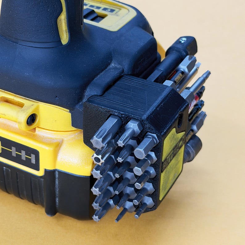 May include: A black 3D-printed bit holder attachment mounted to a yellow and black power drill. The holder contains a grid of various metal screwdriver bits, including star, hex, and flathead tips, arranged in rows for organised storage.