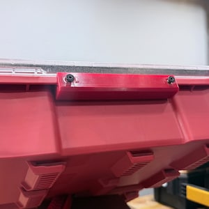 K&ouml;nnte beinhalten: Ein roter Kunststoff-Aufbewahrungsbeh&auml;lter mit einem transparenten Deckel und einem rechteckigen roten Griffaufsatz, der mit zwei schwarzen Schrauben befestigt ist. Der Beh&auml;lter hat eine geformte, texturierte Au&szlig;enfl&auml;che mit sichtbaren Rippen.
