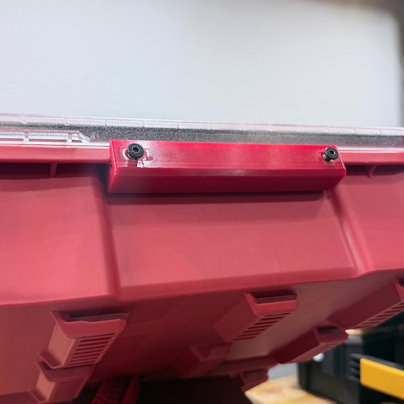 K&ouml;nnte beinhalten: Ein roter Kunststoff-Aufbewahrungsbeh&auml;lter mit einem transparenten Deckel und einem rechteckigen roten Griffaufsatz, der mit zwei schwarzen Schrauben befestigt ist. Der Beh&auml;lter hat eine geformte, texturierte Au&szlig;enfl&auml;che mit sichtbaren Rippen.