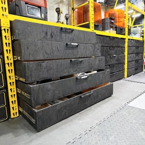 Może przedstawiać: Szereg szerokich, czarnych drewnianych szuflad do przechowywania zainstalowanych w żółtym metalowym regale przemysłowym. Szuflady posiadają czarne poziome uchwyty i metalowe prowadnice kulkowe, a niektóre z nich są częściowo wysunięte.