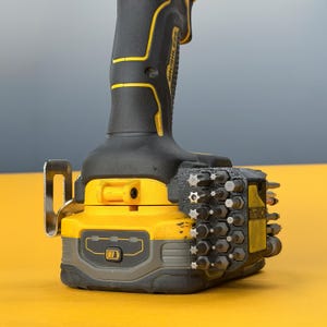 May include: A yellow and black cordless power drill featuring a brushless motor label on the handle, a metal belt hook, and a black plastic bit holder attachment mounted to the side, containing multiple steel screwdriver bits of various sizes.