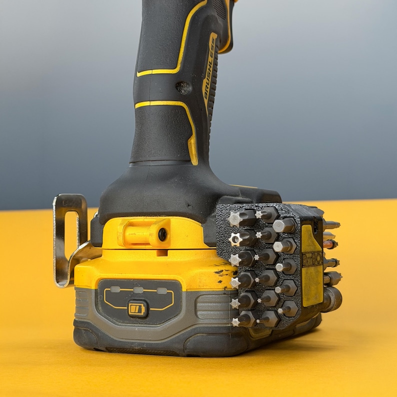 May include: A yellow and black cordless power drill featuring a brushless motor label on the handle, a metal belt hook, and a black plastic bit holder attachment mounted to the side, containing multiple steel screwdriver bits of various sizes.