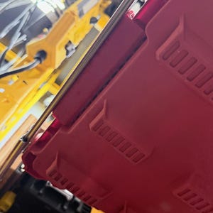 K&ouml;nnte beinhalten: Ein roter Aufbewahrungskoffer aus Kunststoff mit strukturierter Oberfl&auml;che und geformten rechteckigen Vertiefungen. Der Koffer verf&uuml;gt &uuml;ber einen transparenten Deckelbereich und Metallverschl&uuml;sse und ist schr&auml;g &uuml;ber einem gelben Werkzeug.