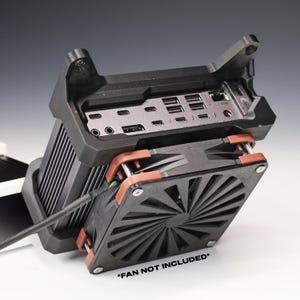 Op de afbeelding: Een zwarte koelventilatorhouder bevestigd aan een metalen elektronisch apparaat met meerdere poorten, waaronder USB-C, HDMI en Ethernet. Het geheel bevat een zwarte ventilator met roodbruine hoekclips en de tekst *FAN NOT INCLUDED*.