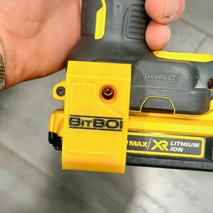 May include: A yellow plastic BitBoi bit holder attachment mounted on a black and yellow DeWalt Max XR Lithium Ion power tool battery. The accessory features a textured surface and a single screw hole, securely fastened to the side of the tool.