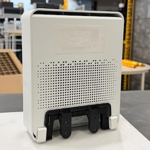 May include: A white rectangular electronic device with rounded corners. The back panel has a grid of small holes and a black mounting bracket. A sticker is affixed to the top of the device. The device is sitting on a white surface.