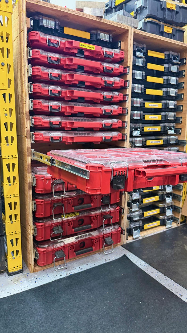 K&ouml;nnte beinhalten: Ein Holzregal mit mehreren roten und schwarzen Kunststoff-Werkzeugkoffern mit transparenten Deckeln. Ein roter Koffer ist auf einer Metallschubladenschiene ausgezogen. Die Einheiten verf&uuml;gen &uuml;ber schwarze Verschl&uuml;sse und gelbe Etiketten.
