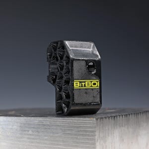 May include: A black 3D-printed component featuring a honeycomb-like geometric side pattern and a flat front face. The front displays the yellow text BITBOI printed on a rectangular background, with a small circular opening positioned above the text.
