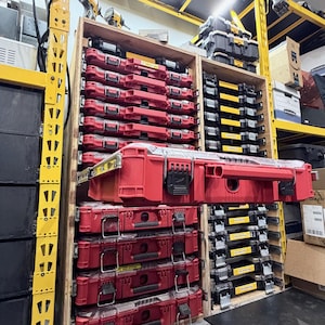 K&ouml;nnte beinhalten: Ein Holzregalsystem mit mehreren vertikal gestapelten roten Kunststoff-Werkzeugkoffern, wobei eine Einheit auf einer Gleitschiene nach au&szlig;en ausgezogen ist. Das Regal befindet sich in einem gelben Metallgestell mit weiteren schwarzen Koffern.