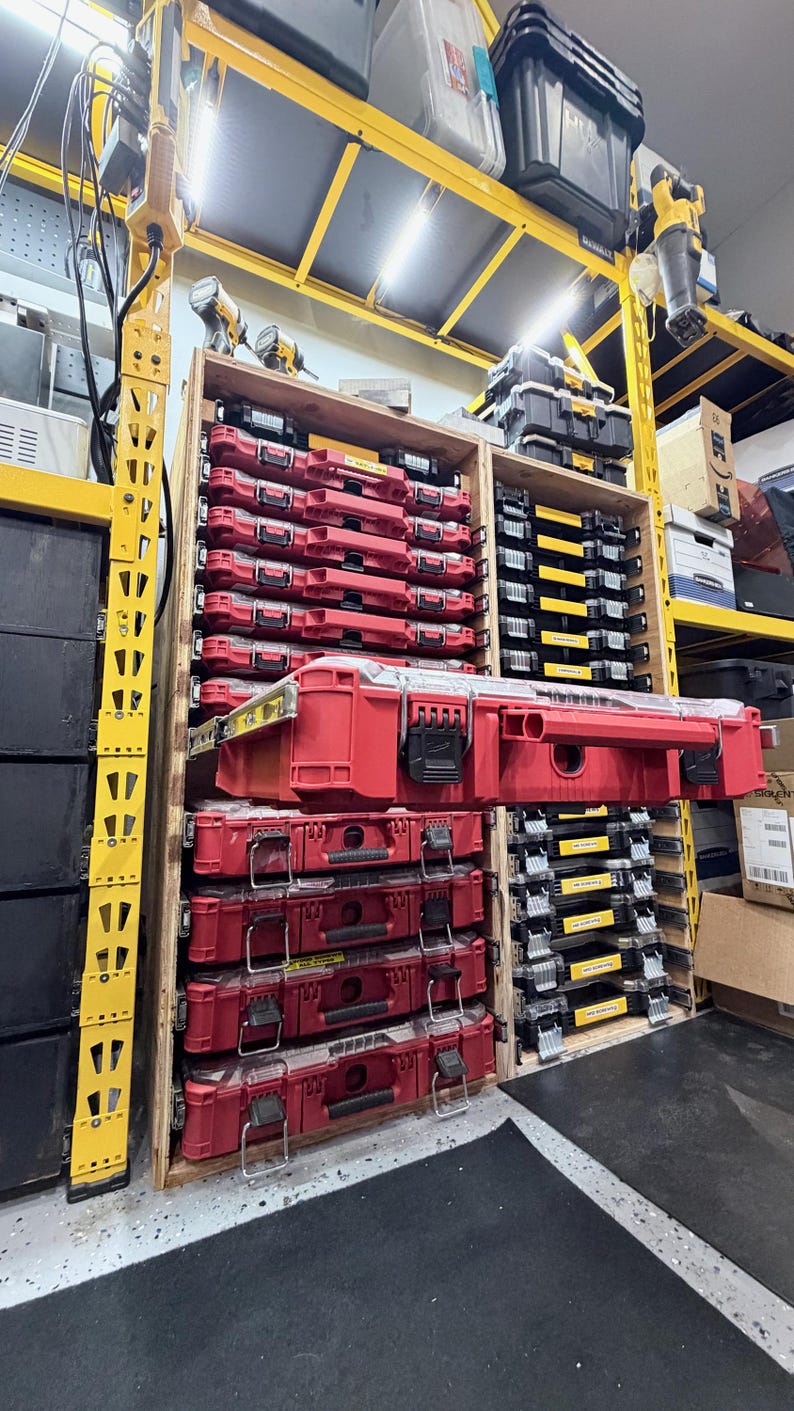 K&ouml;nnte beinhalten: Ein Holzregalsystem mit mehreren vertikal gestapelten roten Kunststoff-Werkzeugkoffern, wobei eine Einheit auf einer Gleitschiene nach au&szlig;en ausgezogen ist. Das Regal befindet sich in einem gelben Metallgestell mit weiteren schwarzen Koffern.
