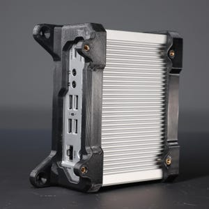 May include: A silver, rectangular electronic device with a ribbed aluminium casing, encased in a black, 3D-printed protective frame. The front panel features multiple USB ports, audio jacks, and a network port, with visible mounting holes.