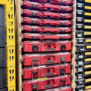 May include: A wooden storage unit filled with red toolboxes. The toolboxes are stacked in rows and have black handles and clear lids. Some toolboxes have labels that read "BATTERIES".
