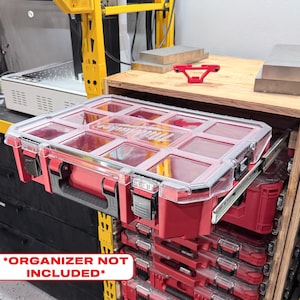 Puede incluir: Un maletín organizador Milwaukee de plástico rojo y transparente con múltiples compartimentos internos, que cuenta con un asa negra y cierres de seguridad, montado sobre un sistema de rieles deslizantes de metal.