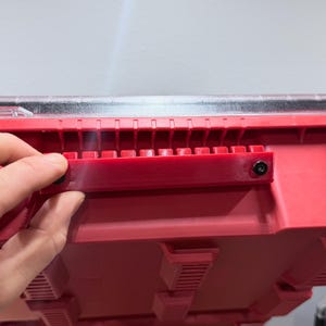 K&ouml;nnte beinhalten: Ein roter Kunststoff-Aufbewahrungsbeh&auml;lter mit einer horizontalen roten Montagehalterung, die mit zwei schwarzen Schrauben befestigt ist. Der Beh&auml;lter hat einen transparenten Deckel und eine Reihe vertikaler Rippen entlang der Oberkante.