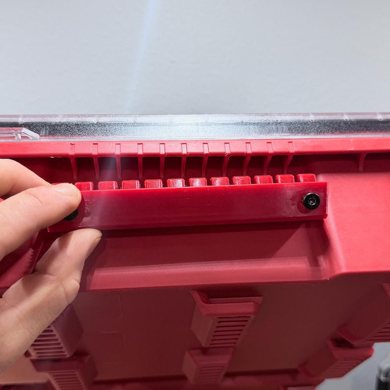 K&ouml;nnte beinhalten: Ein roter Kunststoff-Aufbewahrungsbeh&auml;lter mit einer horizontalen roten Montagehalterung, die mit zwei schwarzen Schrauben befestigt ist. Der Beh&auml;lter hat einen transparenten Deckel und eine Reihe vertikaler Rippen entlang der Oberkante.