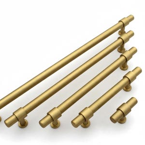 Pode incluir: Um conjunto de puxadores dourados para armários em vários comprimentos. Cada puxador apresenta uma seção central cilíndrica e extremidades arredondadas, montadas em pequenas bases circulares. O hardware está disposto diagonalmente sobre um fundo branco.