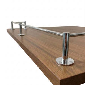 May include: A wooden shelf with a polished, dark brown finish. It features a chrome-colored metal railing attached with circular mounting plates. The shelf is designed for display or storage.