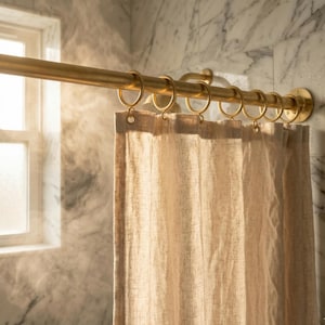 Peut inclure: Une tringle de rideau de douche dorée avec des anneaux assortis et un rideau de douche en lin beige. La tringle est fixée sur un mur en carrelage de marbre. Une fenêtre est visible en arrière-plan.