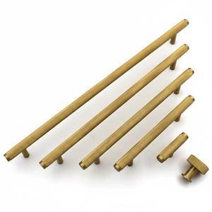 Puede incluir: Un conjunto de seis tiradores y pomos de armario de color latón. Los tiradores tienen forma de barra con una superficie texturizada, de diferentes longitudes. El pomo tiene un diseño texturizado similar. Sobre fondo blanco.