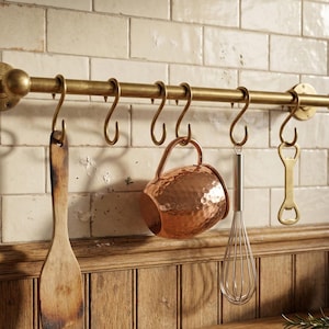 Könnte beinhalten: Eine Küchenreling aus Messing mit hängenden Utensilien und Zubehör. Enthält einen Holzspatel, einen Kupferbecher, einen Schneebesen und einen Flaschenöffner. Die Reling ist an einer gefliesten Wand über einer Holztheke montiert.