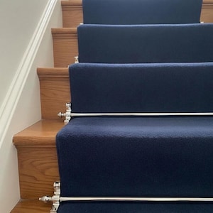 Puede incluir: Una escalera con escalones de madera y alfombras corredizas azul marino. La alfombra está asegurada con barras plateadas y terminales decorativos. Los escalones de madera son de color marrón claro y la pared es blanca.