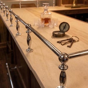 May include: A polished chrome bar rail with decorative supports is installed on a light wood bar. A decanter, glasses, compass, and keys are on the bar. The bar has a dark wood front.