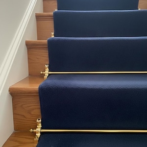 Puede incluir: Una escalera de madera con alfombras corredizas azul marino. La alfombra está asegurada con varillas doradas y soportes decorativos. La pared está pintada de blanco.