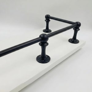 May include: A black metal towel rack with a decorative design, mounted on a white shelf. The rack features a horizontal bar supported by ornate vertical posts with rounded finials. The overall design is classic and elegant.