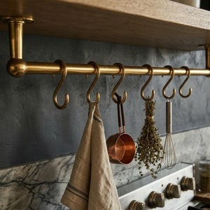 Puede incluir: Un estante de cocina de latón con utensilios y una toalla colgantes. El estante está montado debajo de un estante de madera. Los artículos colgantes incluyen una cacerola de cobre, un manojo de hierbas y un batidor. El fondo presenta una pared gris oscuro y una encimera de mármol.