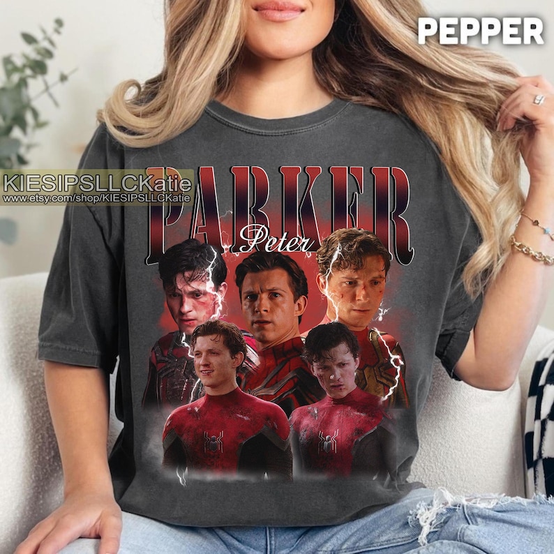 Pu&ograve; includere: T-shirt grigio scuro con una grafica di Peter Parker, il nome "PARKER" in rosso e "Peter" in bianco. La maglietta ha uno stile vintage e la scritta "PEPPER" &egrave; stampata in alto a destra.