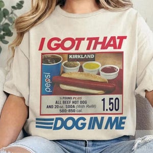 May include: Cream-colored t-shirt with a graphic of a hot dog meal, including a hot dog, condiments, and a Pepsi. The text "I GOT THAT" is above the graphic, and "DOG IN ME" is below. The graphic includes the text "KIRKLAND".
