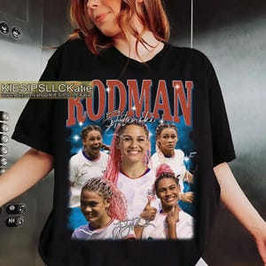 May include: Black t-shirt with a graphic of a soccer player with pink hair, the name "RODMAN" in large red letters, and the name "Stutinski". The shirt also includes the text "KIESIPSLLCKatie" and the Etsy shop URL.