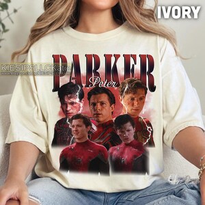Pu&ograve; includere: T-shirt color avorio con grafica di Peter Parker, il nome "PARKER" in rosso e pi&ugrave; immagini del personaggio in tuta rossa e nera. &Egrave; visibile anche il testo "Peter". La maglietta &egrave; di colore crema chiaro.