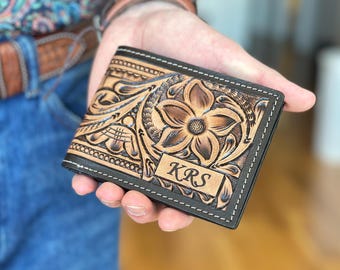 May include: A brown leather wallet with intricate floral tooling and a black border. The wallet is personalized with the initials "KRS".