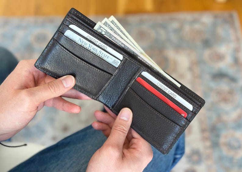 May include: A brown leather wallet with a credit card and cash inside. The wallet is open and the cash is sticking out of the top. The wallet has a flap that closes with a snap.