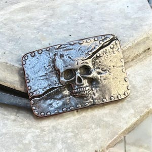 May include: A rectangular silver-colored belt buckle with a detailed skull design. The skull is centered on the buckle, with visible teeth and eye sockets. The buckle has a textured surface and is edged with small rivets.
