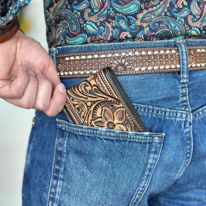 May include: A brown leather wallet with a floral design is tucked into the back pocket of a pair of blue jeans.