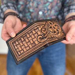 May include: A brown leather wallet with a tooled design featuring a floral pattern and a checkered background. The wallet has a black border and stitching.