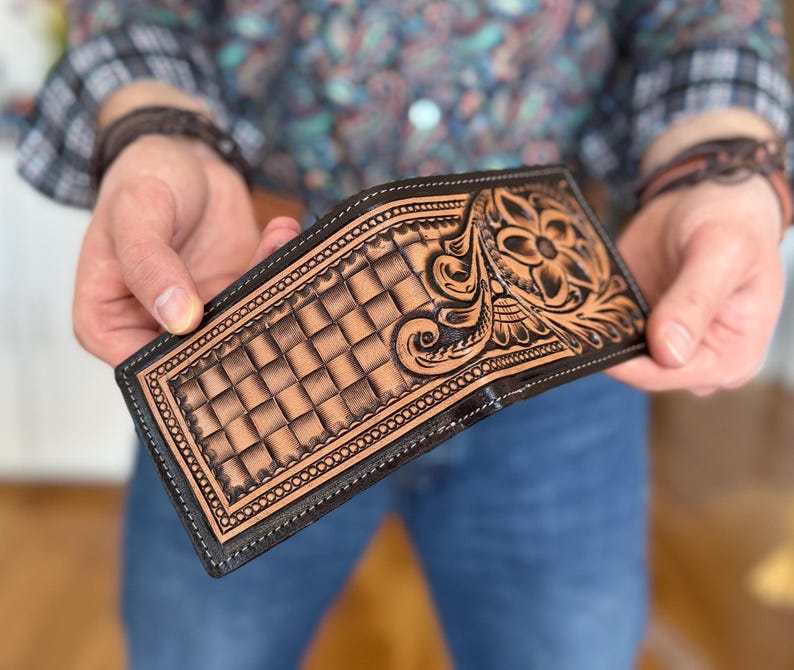 May include: A brown leather wallet with a tooled design featuring a floral pattern and a checkered background. The wallet has a black border and stitching.
