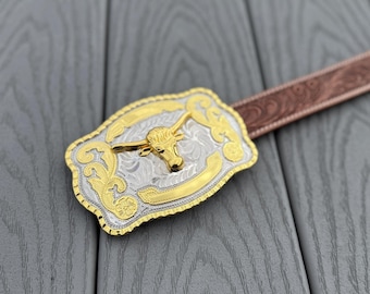 May include: A gold and silver-toned belt buckle with a longhorn design. The buckle has an ornate, engraved pattern and is attached to a brown leather belt. The buckle is rectangular with rounded edges.