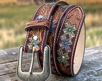 May include: A dark brown leather belt featuring intricate floral tooling with hand-painted turquoise and blue accents. The belt includes a silver-toned metal buckle with engraved scrollwork and white contrast stitching along the edges.