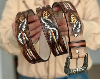May include: A brown leather belt featuring an embossed arrow pattern with white and gold accents. The belt includes a silver-toned metal buckle with intricate scrollwork engravings and two metal rivets securing the strap near the buckle attachment.