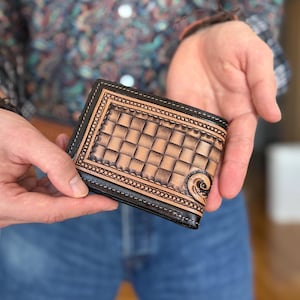 May include: A brown leather wallet with a tooled design and a black border. The wallet is closed and has a stitched edge.