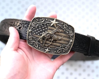 May include: A black leather belt featuring an intricate embossed floral pattern and a rectangular brass-toned belt buckle. The buckle displays a raised relief of a bucking bronco horse and rider set against a background of the American flag.