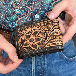 May include: A brown leather wallet with intricate floral tooling. The wallet has a dark brown border and stitching.