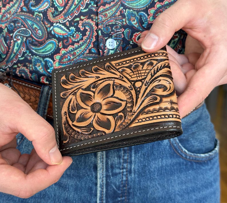 May include: A brown leather wallet with intricate floral tooling. The wallet has a dark brown border and stitching.