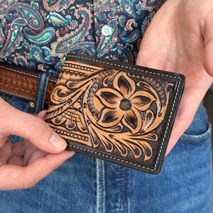 May include: A brown leather wallet with intricate floral tooling. The wallet has a black border and stitching.