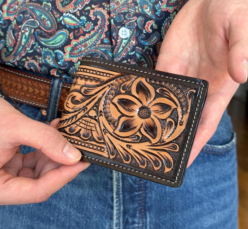 May include: A brown leather wallet with intricate floral tooling. The wallet has a black border and stitching.