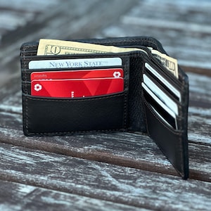 May include: A brown leather wallet with multiple card slots and a billfold. The wallet is open and contains several credit cards, including one with the text "New York State USA" and a red credit card with a recycling symbol. There are also several bills in the billfold.