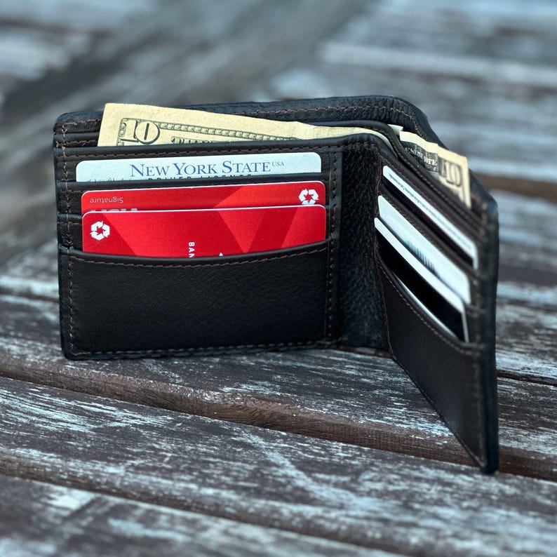 May include: A brown leather wallet with multiple card slots and a billfold. The wallet is open and contains several credit cards, including one with the text "New York State USA" and a red credit card with a recycling symbol. There are also several bills in the billfold.